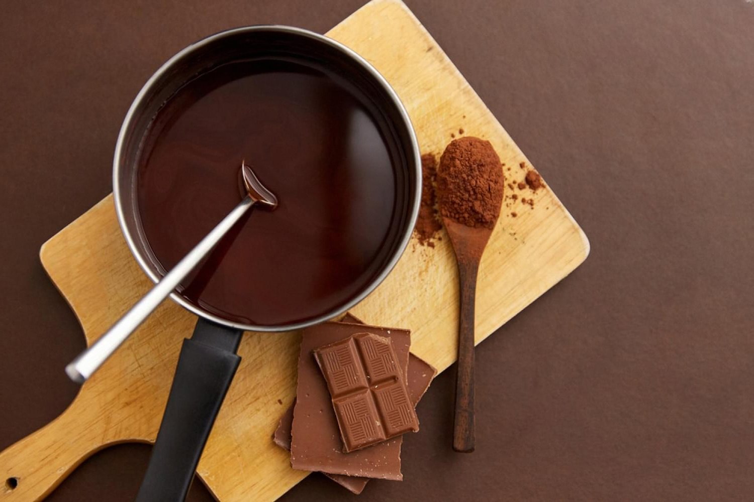 Melted Belgian chocolate in a saucepan next to milk chocolate squares and a spoon of cocoa powder